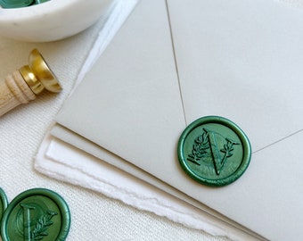 May include: A green wax seal with a leafy design and the letter "V" in the center. The seal is on a white envelope.
