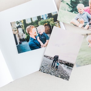 May include: A photo album with several photos of a father and son. The photos show the father holding the son, the father and son walking on a beach, and the father and son sitting on a grassy hill.