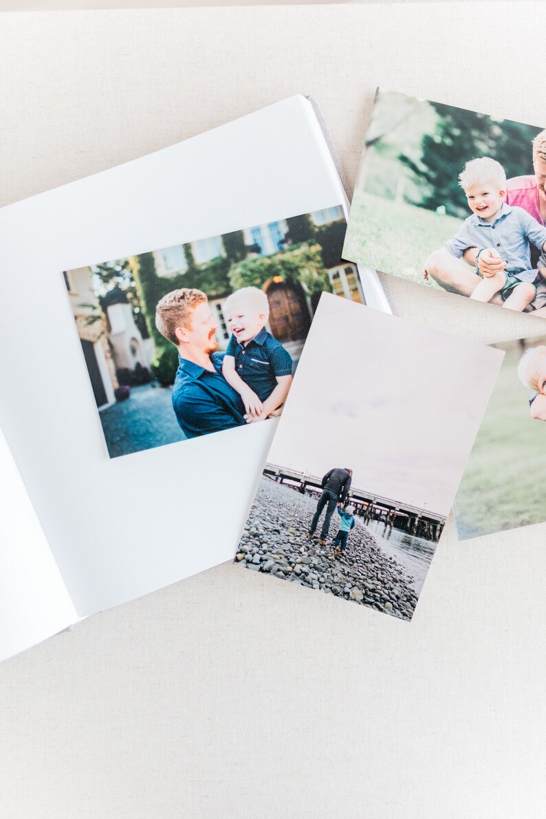 May include: A photo album with several photos of a father and son. The photos show the father holding the son, the father and son walking on a beach, and the father and son sitting on a grassy hill.