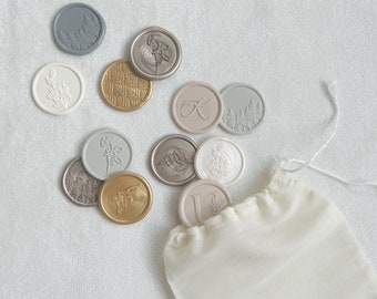 Può includere: Una collezione di sigilli di cera in vari colori e design, tra cui motivi floreali, una lettera K e alberi. I sigilli sono in un piccolo sacchetto di stoffa bianco con coulisse.