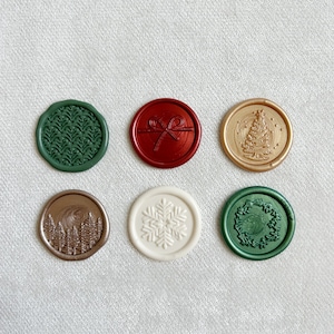 May include: Six round wax seals with different holiday designs. The seals are in various colors, including green, red, gold, and white. The designs include a snowflake, a Christmas tree, a wreath, a bow, a forest, and a pine tree.