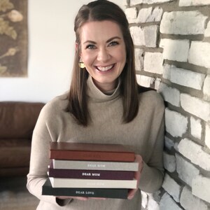 May include: A woman is holding a stack of five small, hardcover books. The books are all different colors and have the words "Dear Mom" and "Dear Love" printed on the covers.