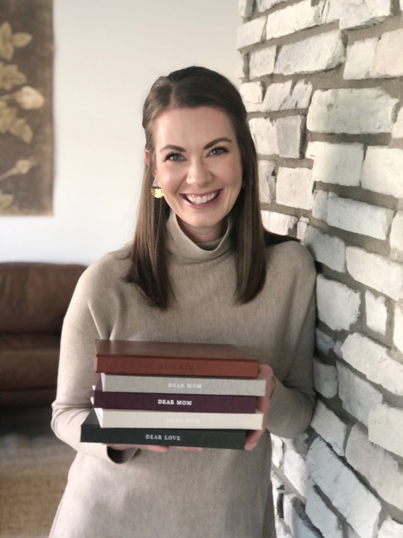May include: A woman is holding a stack of five small, hardcover books. The books are all different colors and have the words "Dear Mom" and "Dear Love" printed on the covers.