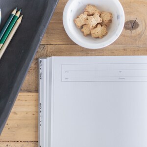 May include: A blank, white journal with a black spine and a black tray of colored pencils. The journal has lines for "From", "Day", "Age", and "Occasion".