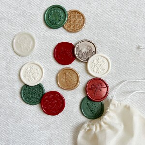 May include: A collection of 12 assorted wax seals in a variety of colors and designs, including snowflakes, trees, wreaths, and a bow. The seals are arranged on a white surface with a small white bag in the lower right corner.