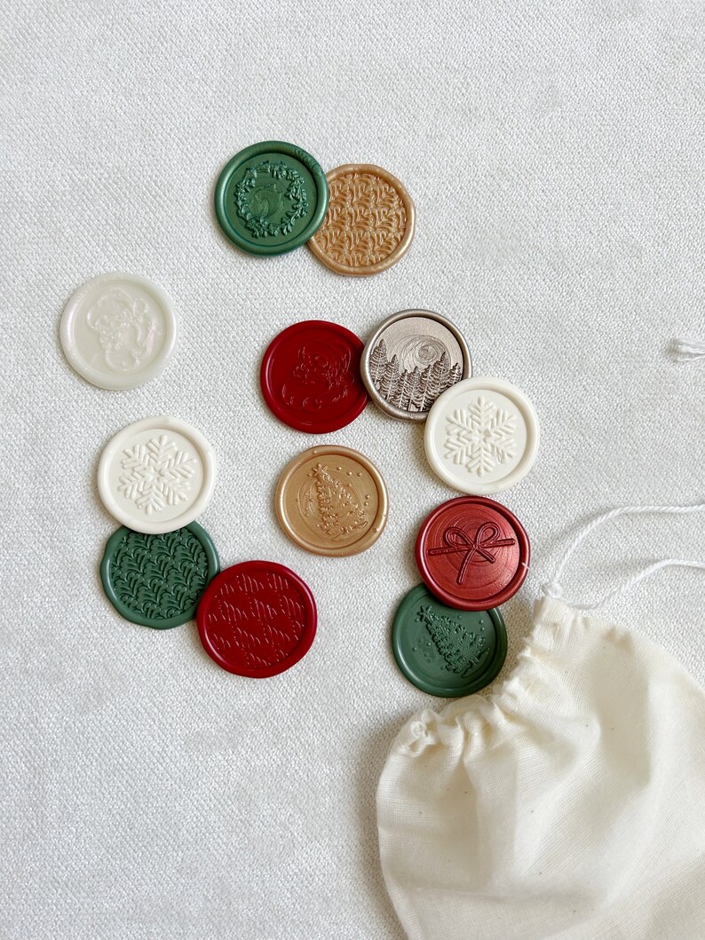 May include: A collection of 12 assorted wax seals in a variety of colors and designs, including snowflakes, trees, wreaths, and a bow. The seals are arranged on a white surface with a small white bag in the lower right corner.