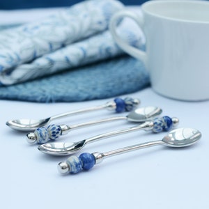 May include: Four silver teaspoons with blue and white ceramic handles. The spoons are arranged in a row on a white surface.