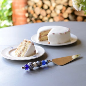 Könnte beinhalten: Ein weißer Kuchen mit weißem Frosting und rosa Dekorationen steht auf einem weißen Teller. Ein Stück Kuchen liegt auf einem blau-weiß gemusterten Teller. Ein blaues und weißes Perlen-Kuchenmesser liegt auf dem Tisch.