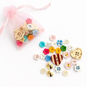 Puede incluir: Una colección de pequeños botones variados en diversas formas, incluyendo flores, corazones y patos, con colores como rosa, azul, amarillo, verde y marrón. Algunos botones son sólidos, mientras que otros tienen patrones de rayas o flores.