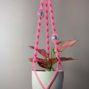 May include: A pink macrame plant hanger featuring knotted cord construction and three small reflective disco ball beads integrated into the vertical strands. The hanger supports a solid light-coloured ceramic pot containing a plant with pink and green leaves.