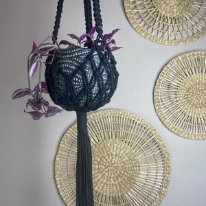 May include: A black macrame plant hanger featuring a knotted net design and a long tassel at the bottom. It holds a dark grey ceramic pot with a textured white pattern, containing a trailing plant with purple and green variegated leaves.