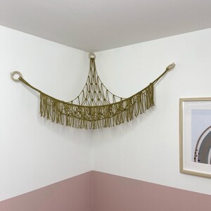 May include: An olive green macrame wall hanging, mounted in a corner on a white wall. The design includes a central net, a fringed bottom, and wooden rings. A framed picture with a rainbow design is also visible on the wall.