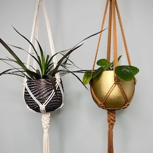 May include: Two hanging planters with macrame cords. The left planter is black with a textured, wavy pattern and white cord. The right planter is metallic gold with a smooth finish and tan cord. Each holds a green plant with distinct foliage.