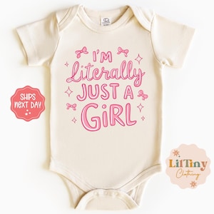 May include: Cream-colored baby bodysuit with the text "I'm Literally Just a Girl" in pink script. The design includes pink bows and stars. A small logo is in the bottom right corner.