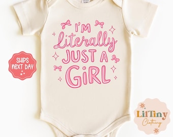 May include: Cream-colored baby bodysuit with the text "I'm Literally Just a Girl" in pink script. The design includes pink bows and stars. A small logo is in the bottom right corner.