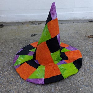 Op de afbeelding: Een kleurrijke patchwork heksenhoed met een brede rand. De hoed heeft een kegelvormige kroon en een cirkelvormige rand, met een levendige mix van oranje, groene, paarse en zwarte stoffen. De hoed is ontworpen voor Halloween of kostuumgebruik.
