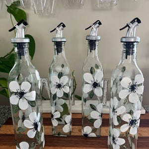 Puede incluir: Cuatro botellas de vidrio transparente con vertedores de color plateado, cada una pintada a mano con flores blancas de centros oscuros y hojas verdes. Las botellas tienen una base cuadrada y están alineadas sobre una superficie de madera.
