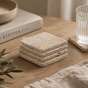 May include: A stack of four square, beige stone coasters with a textured surface sits on a wooden table. A glass of water and a book titled "THE KINFOLK HOME" are also visible. The coasters are designed to protect surfaces from water rings.