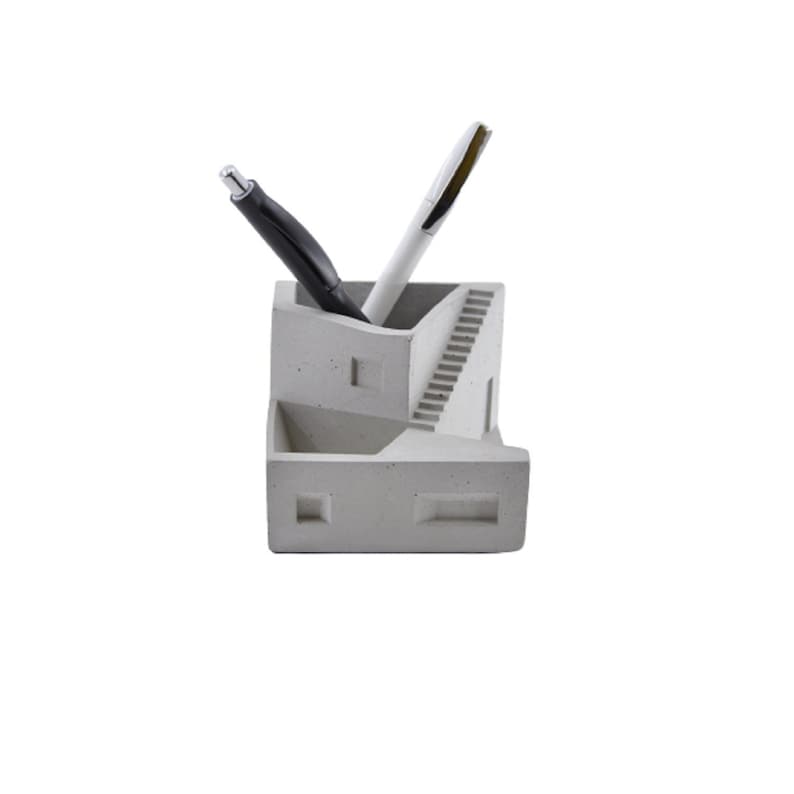 May include: A gray concrete desk organizer shaped like a small house with a staircase. Two pens are in the organizer, one black and one white.