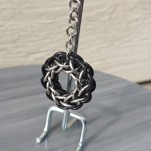 May include: A black and silver chainmail keyring with a circular design. The keyring is attached to a silver metal hook.