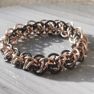May include: A black and rose gold chainmail bracelet. The bracelet is made of small metal rings that are interlocked to create a chainmail pattern.