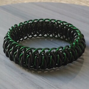 May include: A black and green chainmail bracelet. The bracelet is made of small metal rings that are interlocked to create a flexible and durable piece of jewellery.