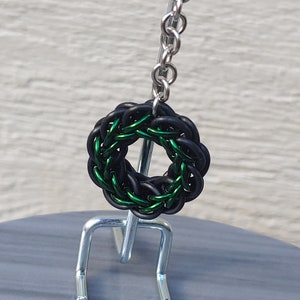 May include: A black and green metal chainmail wreath charm hanging from a silver chain.