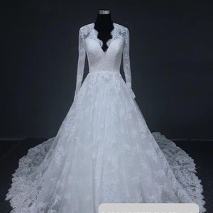 Puede incluir: Un vestido de novia de encaje blanco con escote en V, mangas largas y falda completa. El vestido está en un maniquí y tiene un ribete de encaje. El texto "MUGUET BRIDAL" es visible en la parte inferior de la imagen.