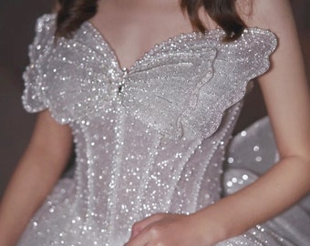 May include: A white, sparkly wedding dress with a sweetheart neckline and a fitted bodice. The dress has a large, flowing skirt and a bow detail on the bodice. The text "MUGUET BRIDAL" is printed on the skirt.