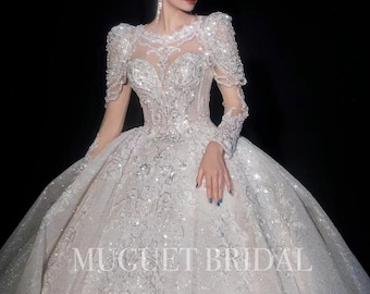 Puede incluir: Un vestido de novia blanco con una falda larga y fluida y un corpiño ajustado. El vestido está cubierto de lentejuelas y cuentas brillantes. El vestido tiene mangas largas y un escote alto. El vestido lo lleva una modelo con una tiara y pendientes. El vestido está etiquetado con "MUGUET BRIDAL".