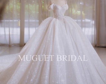 May include: A white wedding dress with a sweetheart neckline and off-the-shoulder straps. The dress is made of a sparkly fabric and has a full skirt. The dress is on a mannequin in a room with white curtains. The text "MUGUET BRIDAL" is printed on the skirt of the dress.