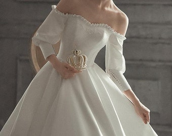 Könnte beinhalten: Ein weißes, schulterfreies Brautkleid mit einem taillierten Oberteil und einem vollem Rock. Das Kleid hat Puffärmel mit Spitzendetails an den Ärmelbündchen. Das Model hält eine goldene Krone.