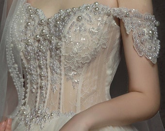 May include: A white off-the-shoulder wedding dress with a tulle skirt and a bodice adorned with pearl and crystal embellishments.