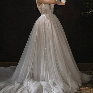 May include: A white tulle wedding dress with a fitted bodice and a flowing skirt. The dress is adorned with delicate sequins and a beaded belt.