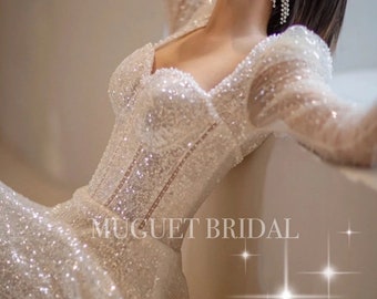 May include: A white, sparkly, long-sleeved wedding dress with a sweetheart neckline and a fitted bodice. The dress has a flowing skirt and is adorned with sequins. The text "MUGUET BRIDAL" is visible on the dress.