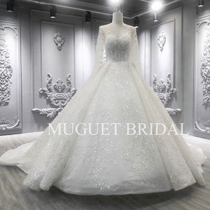 May include: A white wedding dress with long sleeves and a full skirt. The dress is covered in sequins and has a scoop neckline. The dress is photographed in a studio setting with a gray background. The text "MUGUET BRIDAL" is visible on the dress.