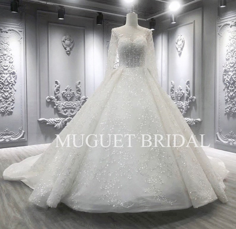 May include: A white wedding dress with long sleeves and a full skirt. The dress is covered in sequins and has a scoop neckline. The dress is photographed in a studio setting with a gray background. The text "MUGUET BRIDAL" is visible on the dress.