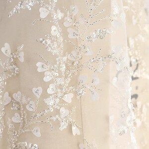 May include: White tulle fabric with a delicate floral pattern made of sequins and beads. The fabric has a soft, flowing texture and is perfect for adding a touch of elegance to any project.
