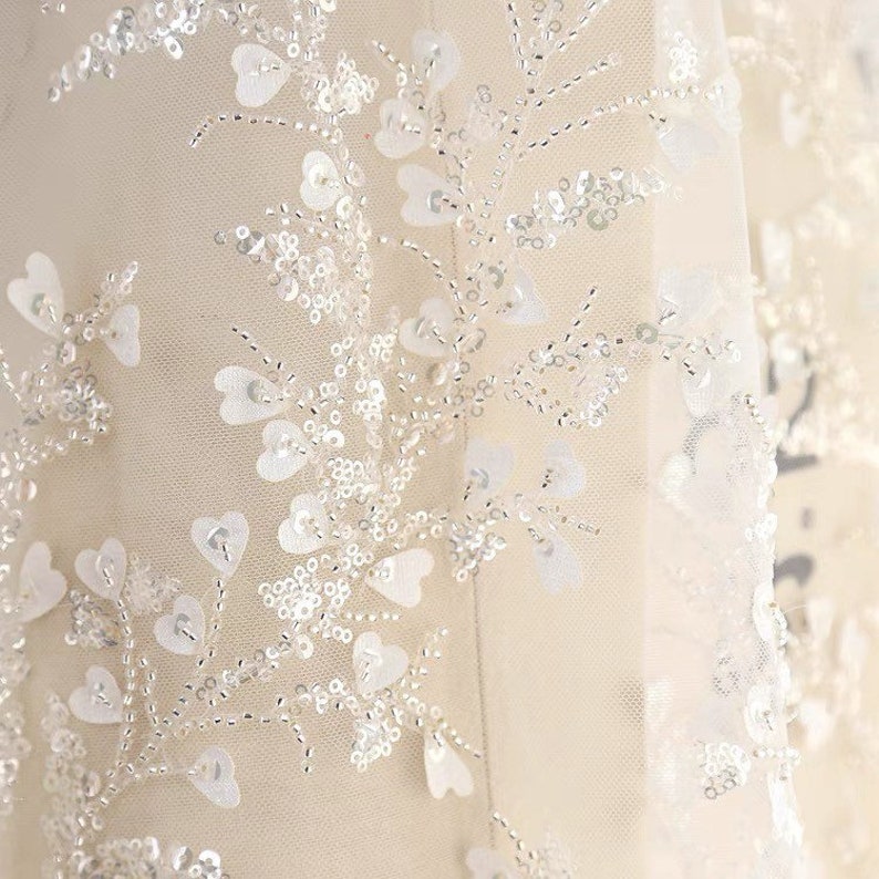 May include: White tulle fabric with a delicate floral pattern made of sequins and beads. The fabric has a soft, flowing texture and is perfect for adding a touch of elegance to any project.