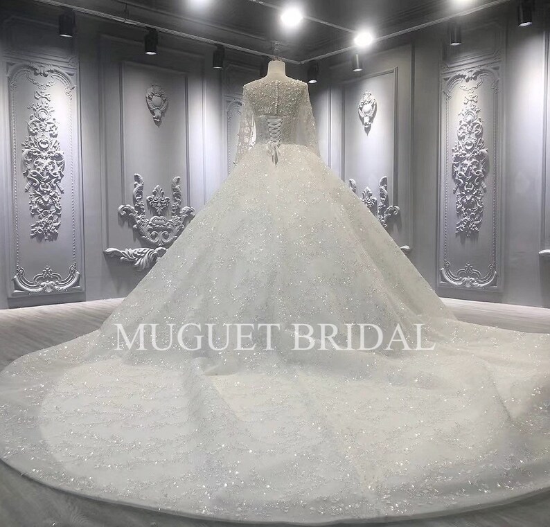 May include: A white wedding dress with a long train and a lace bodice. The dress is covered in sequins and has a fitted bodice with a sweetheart neckline. The dress is shown from the back and has a zipper closure.