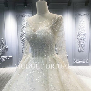 May include: A white wedding dress with long sleeves and a beaded bodice. The dress has a full skirt and is decorated with sequins and beads. The dress is on a mannequin in a store.