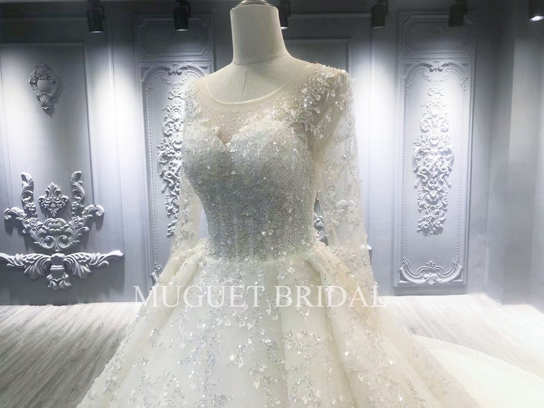 May include: A white wedding dress with long sleeves and a beaded bodice. The dress has a full skirt and is decorated with sequins and beads. The dress is on a mannequin in a store.