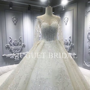 May include: A white wedding dress with long sleeves and a beaded bodice. The dress has a full skirt and a tulle overlay. The dress is displayed on a mannequin in a store setting.