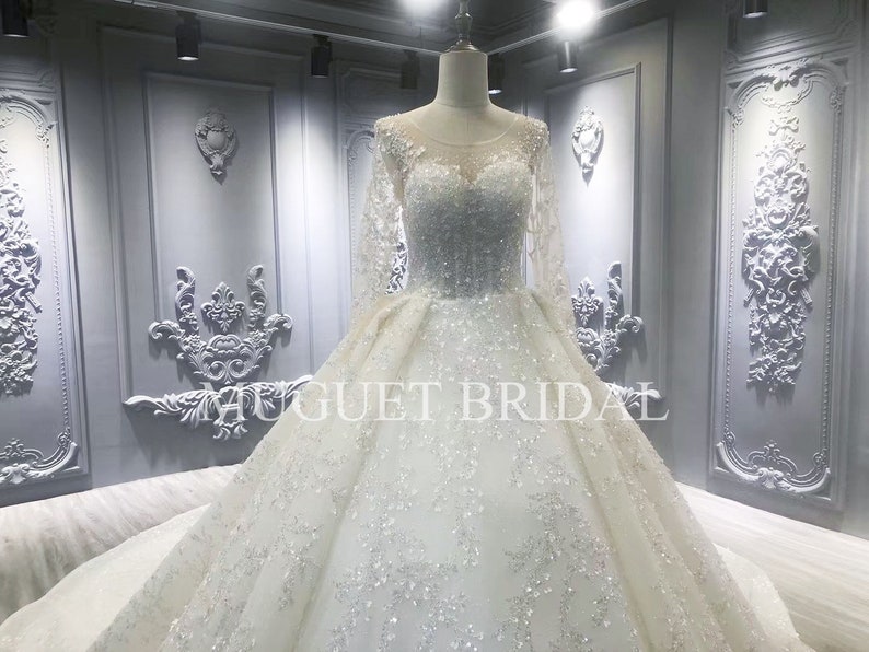 May include: A white wedding dress with long sleeves and a beaded bodice. The dress has a full skirt and a tulle overlay. The dress is displayed on a mannequin in a store setting.