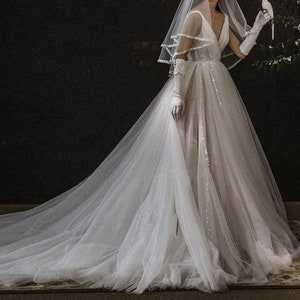 May include: A white tulle wedding dress with a long train and a V-neck bodice. The dress is decorated with sequins and has a sheer veil.
