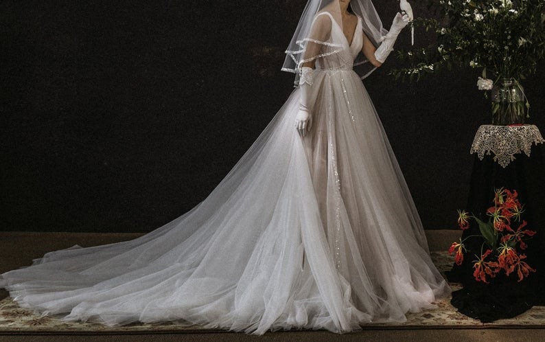 May include: A white tulle wedding dress with a long train and a V-neck bodice. The dress is decorated with sequins and has a sheer veil.