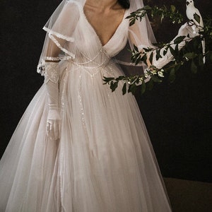 May include: A white tulle wedding dress with a V-neckline and a beaded bodice. The dress has a long train and is adorned with delicate lace details. The model is wearing white gloves and a veil.