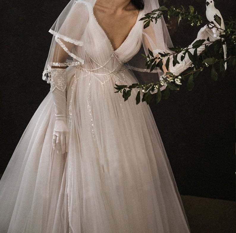 May include: A white tulle wedding dress with a V-neckline and a beaded bodice. The dress has a long train and is adorned with delicate lace details. The model is wearing white gloves and a veil.