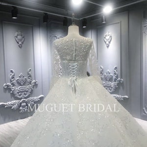 May include: A white wedding dress with long sleeves and a beaded bodice. The dress has a full skirt and a lace-up back. The dress is displayed in a room with white walls and ornate moldings. The text "MUGUET BRIDAL" is visible on the skirt.