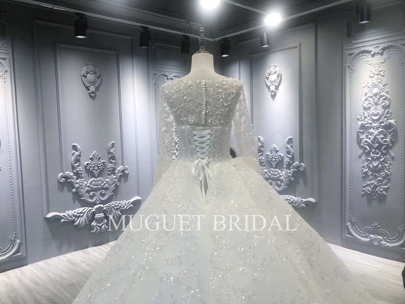 May include: A white wedding dress with long sleeves and a beaded bodice. The dress has a full skirt and a lace-up back. The dress is displayed in a room with white walls and ornate moldings. The text "MUGUET BRIDAL" is visible on the skirt.